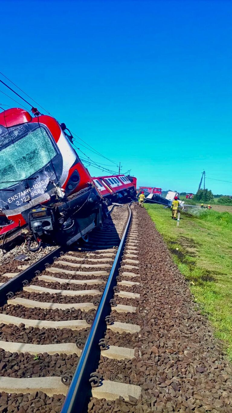Wykolejony, czerwono-czarny pociąg pasażerski z rozbitą szybą. W tle strażacy i zniszczona ciężarówka na przejeździe.