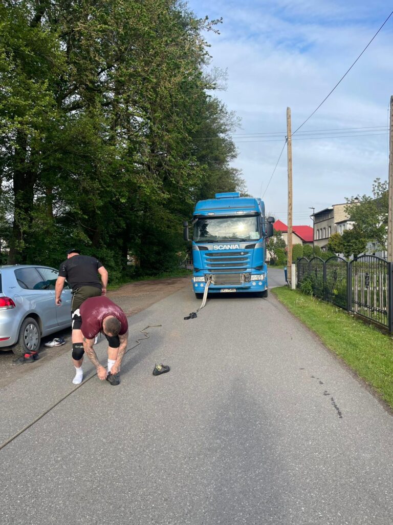 Dwóch mężczyzn na drodze przygotowuje się do przeciągania niebieskiej ciężarówki marki Scania za pomocą grubej liny.